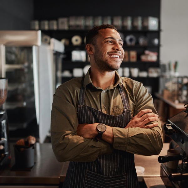 một nam thanh niên mỉm cười, mặc tạp dề, đứng cạnh máy pha espresso trong một quán cà phê độc lập